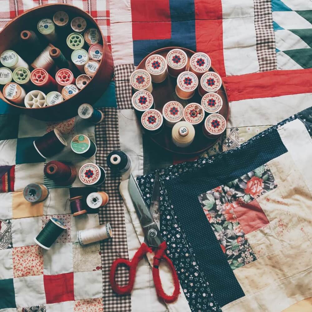 A patchwork quilt with thread spools, needles, and scissors arranged on top. - Home Instead