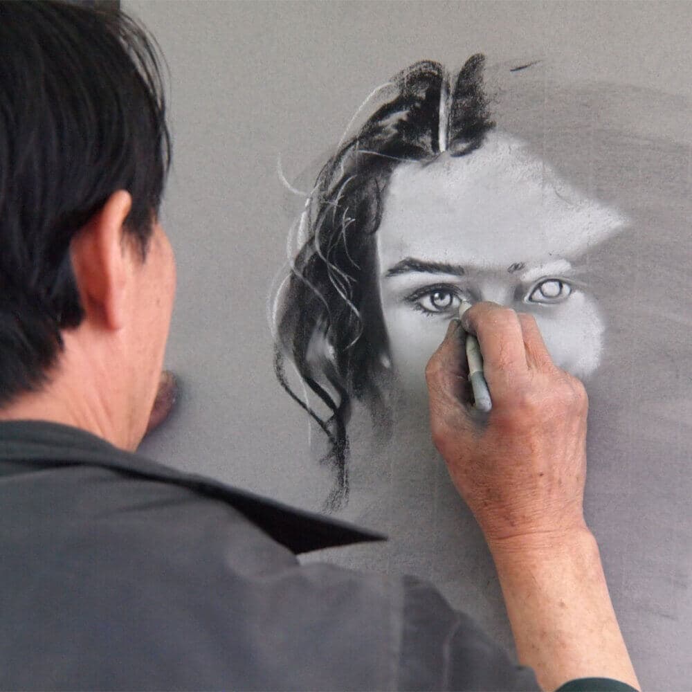 An artist is drawing a woman's face with charcoal on a gray canvas, focusing on detailing the eyes and hair. - Home Instead