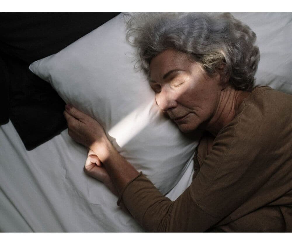 Elderly woman with gray hair sleeping peacefully on white pillow, a beam of light illuminating her face. - Home Instead