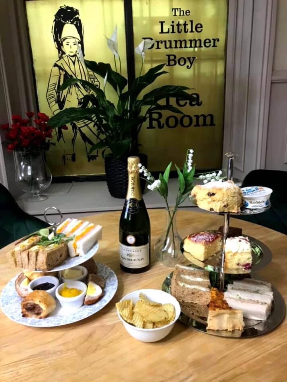 A table with a tiered stand of sandwiches and desserts, champagne, and crisps in "The Little Drummer Boy Tea Room". - Home Instead