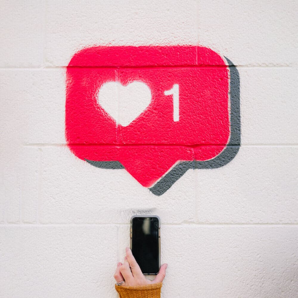Hand holding a smartphone takes a picture of a red 'like' icon spray-painted on a white brick wall. - Home Instead