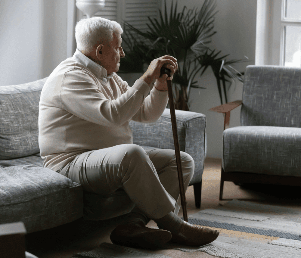 Elderly man with white hair sitting on a gray couch, holding a wooden cane, in a room with plants and furniture. - Home Instead