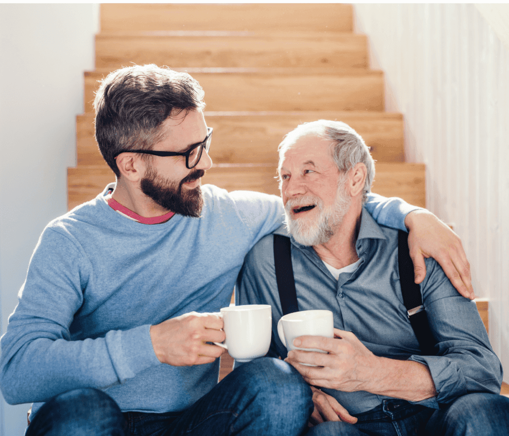 Two men, one older and one younger, sitting on stairs with coffee mugs, smiling and embracing each other warmly. - Home Instead