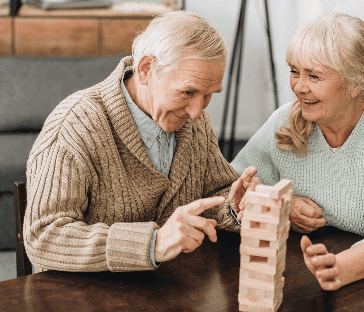An elderly man and woman smiling and playing a game of Jenga together at a wooden table. - Home Instead