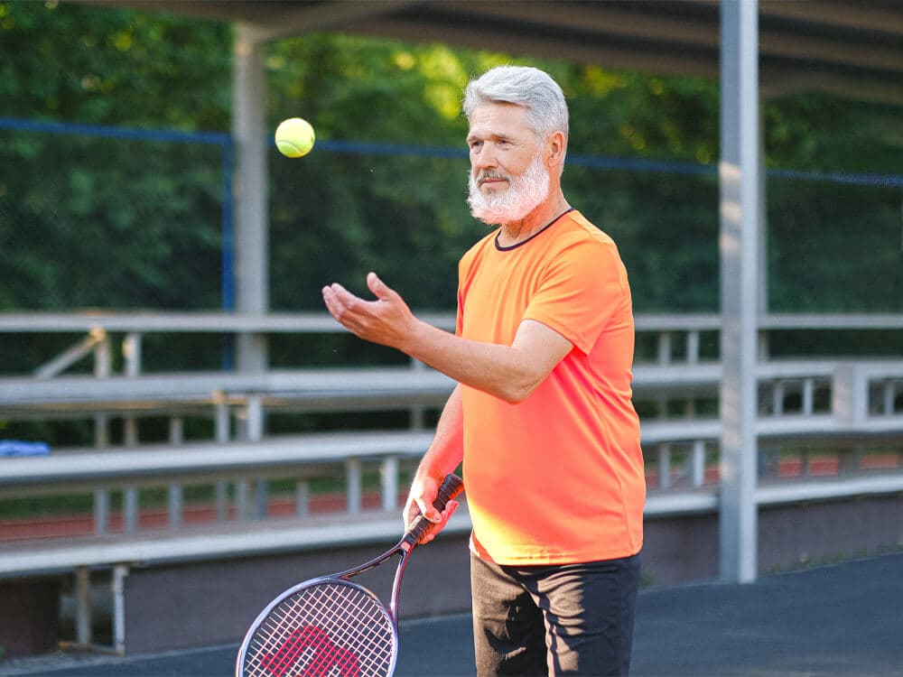 A man with gray hair and a beard, wearing an orange shirt, tossing a tennis ball on a court. - Home Instead