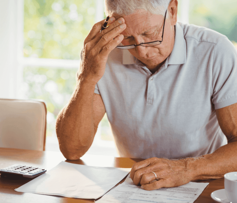 An older man in glasses looks stressed while reviewing documents at a table with a calculator and papers. - Home Instead