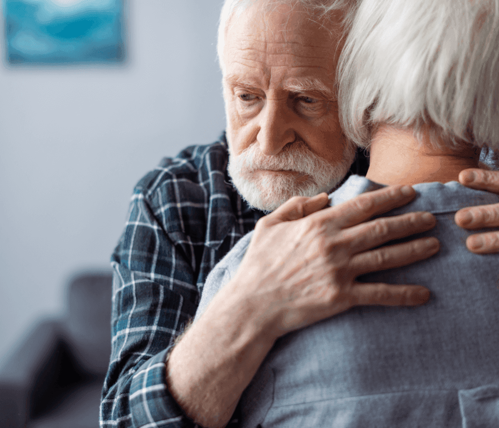 An elderly couple embraces, the man looking concerned while holding the woman who faces away from the camera. - Home Instead
