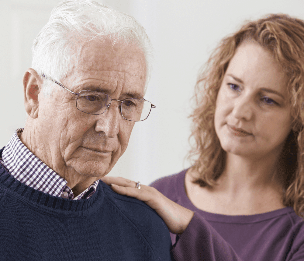 An older man with glasses looks sad while a woman with curly hair offers comfort, placing her hand on his shoulder. - Home Instead