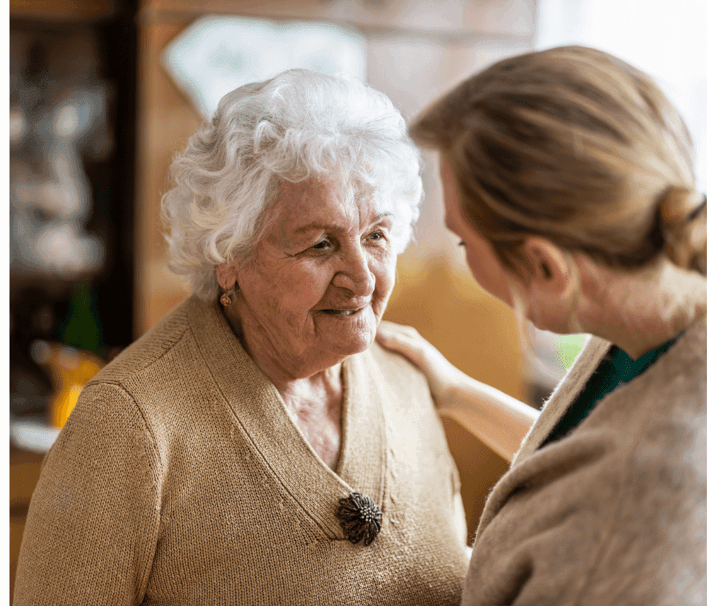 An elderly woman with white hair smiles while talking to a younger woman who is touching her shoulder gently. - Home Instead