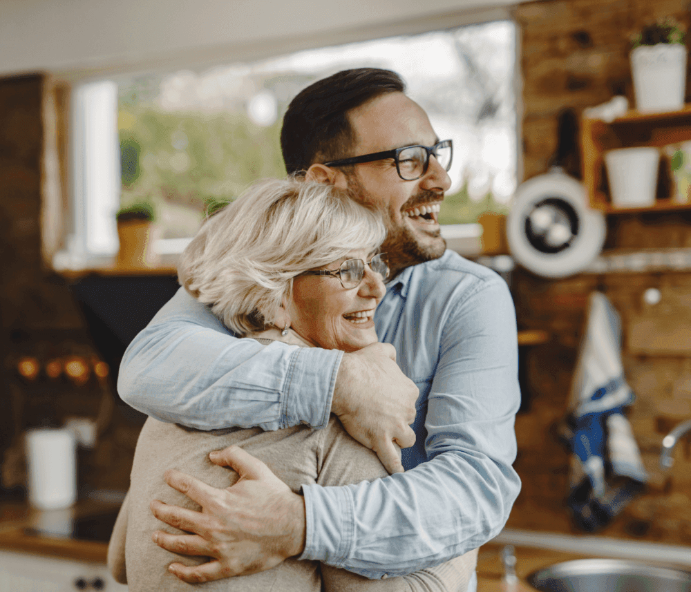 A man and an older woman smiling and hugging in a cozy kitchen. They both wear glasses and seem happy. - Home Instead