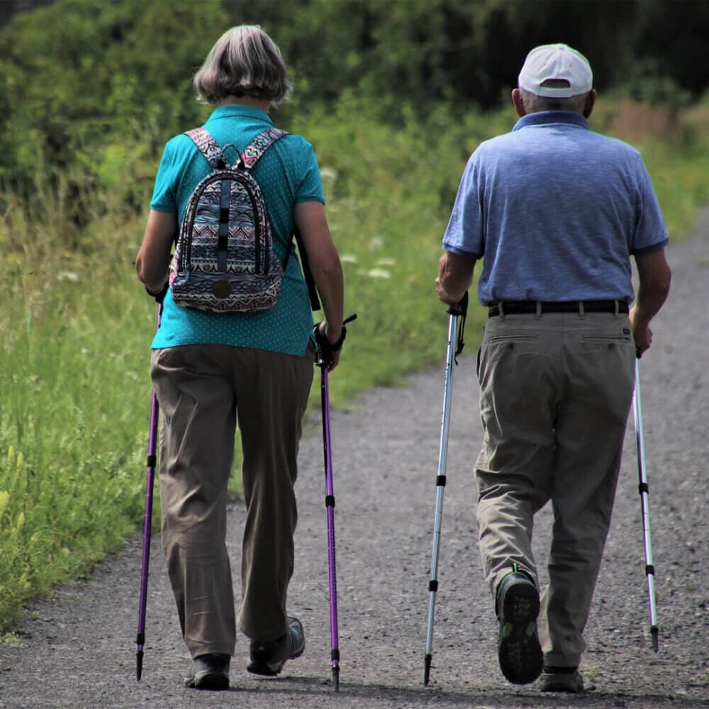 Two elderly people walking with Nordic walking sticks on a gravel path, wearing casual clothing and a backpack. - Home Instead