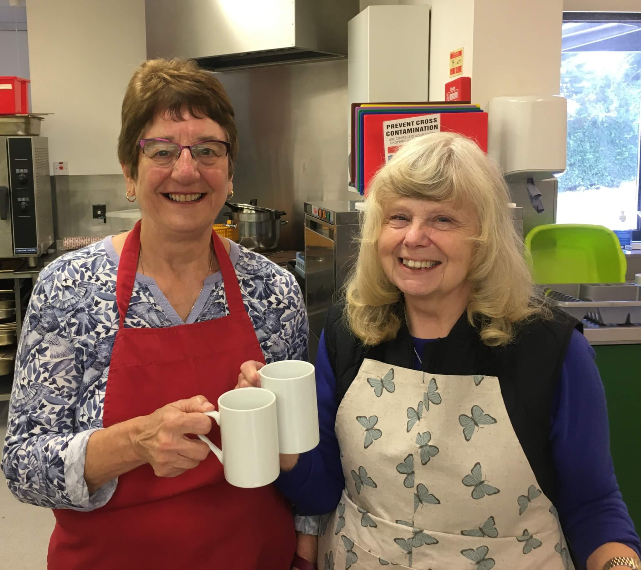 Two women in aprons smiling and holding white mugs in a kitchen. - Home Instead