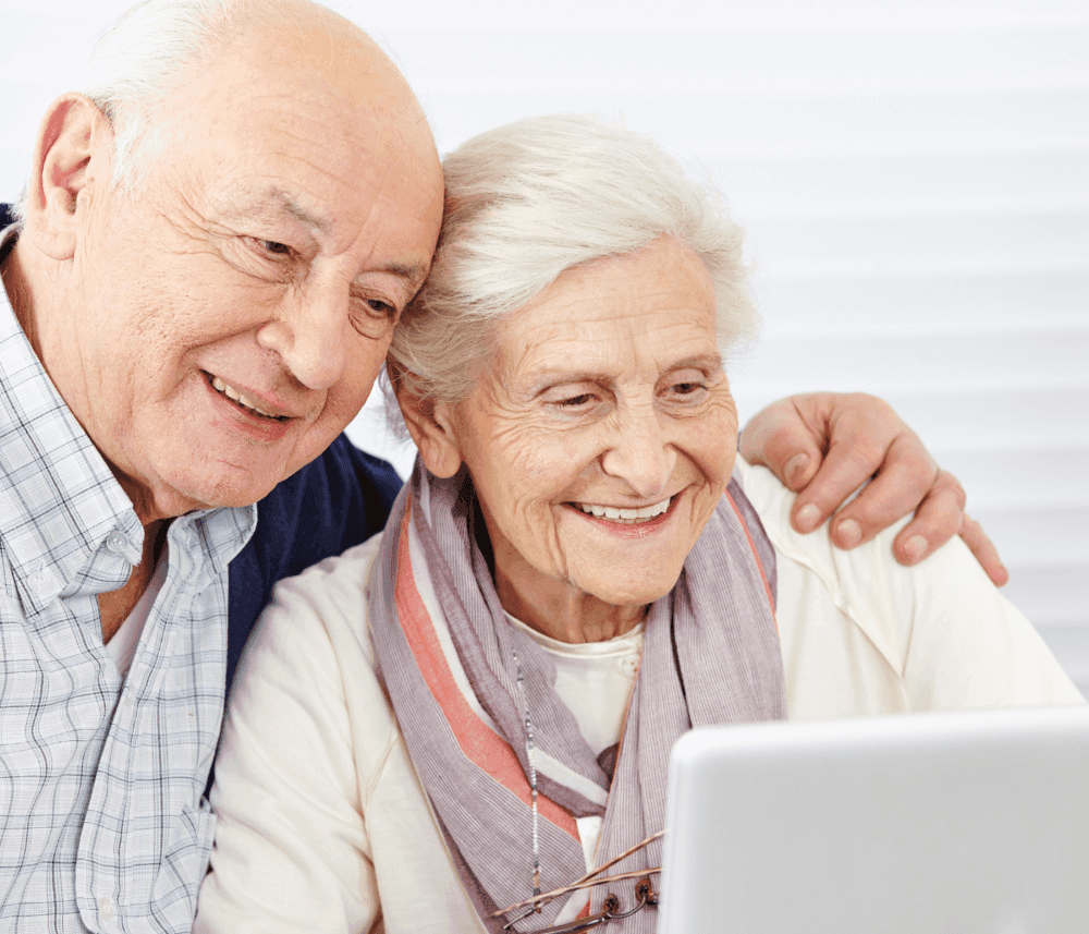 An elderly couple smiling while looking at a laptop screen together, with the man’s arm around the woman. - Home Instead