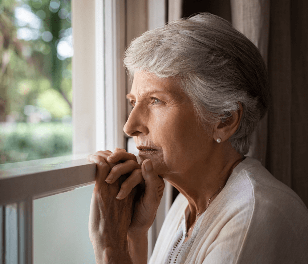Older woman with short gray hair gazes thoughtfully out a window, with her hands gently resting on the window frame. - Home Instead