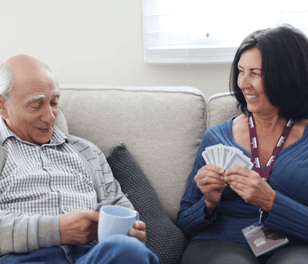 Elderly man and woman smiling while sitting on a couch, the woman holding cards and wearing a name badge. - Home Instead