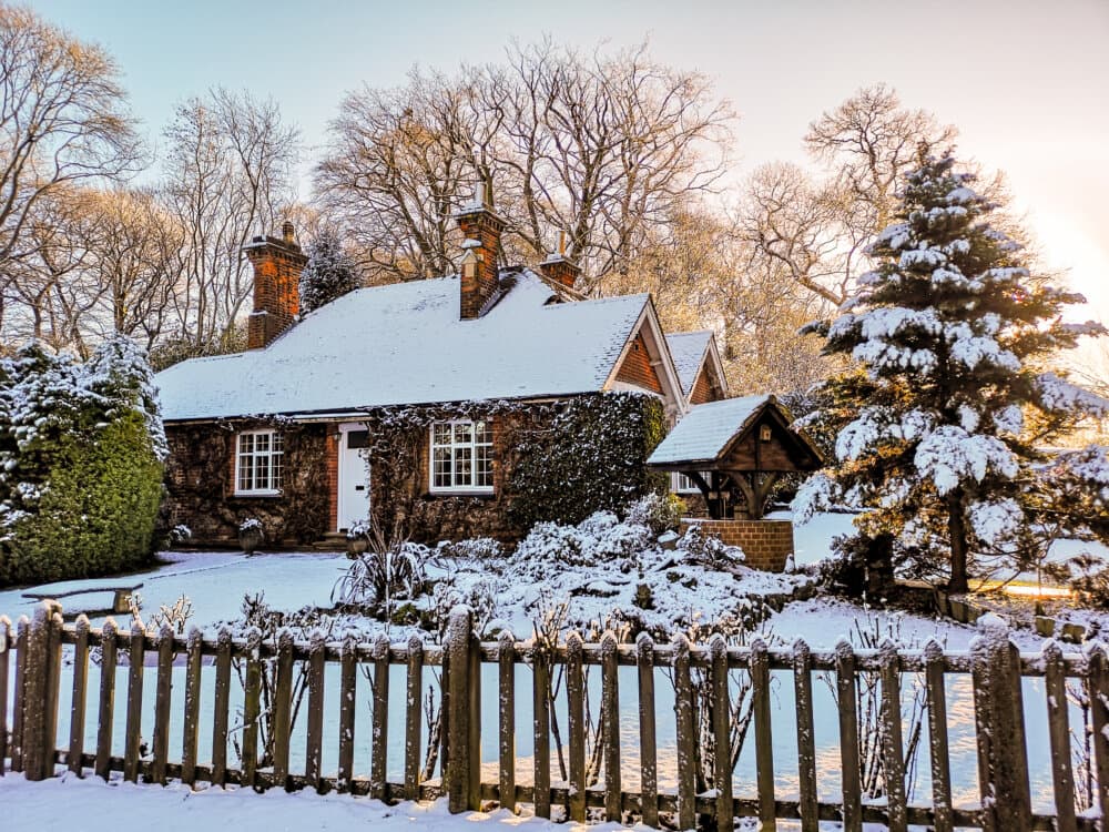 Snow-covered cottage surrounded by trees and a wooden fence, with sunlight filtering through the winter landscape. - Home Instead
