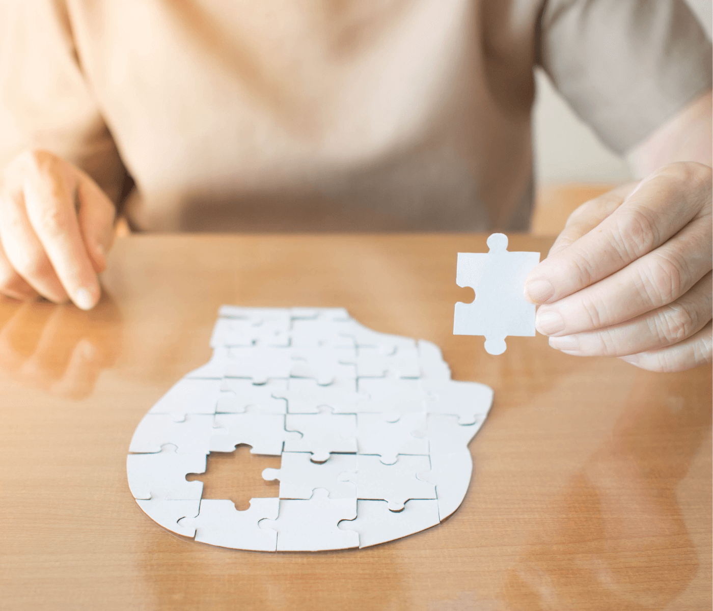 A person assembles a white jigsaw puzzle shaped like a human head on a wooden table, holding a piece in their hand. - Home Instead
