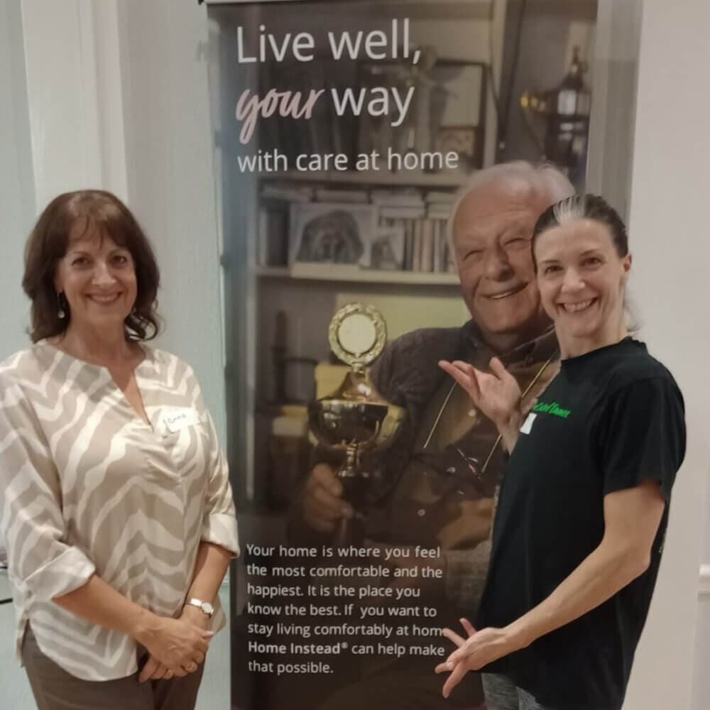 Two women smiling in front of a Home Instead banner promoting senior care services. - Home Instead