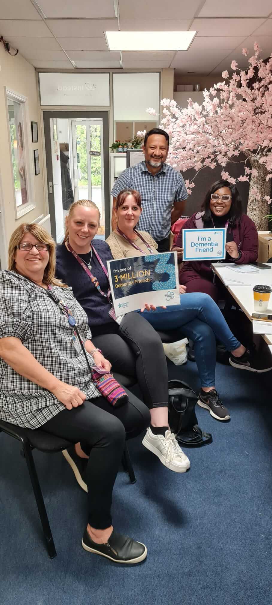Group of five people smiling, holding a poster about supporting people living with dementia, in an office setting. - Home Instead