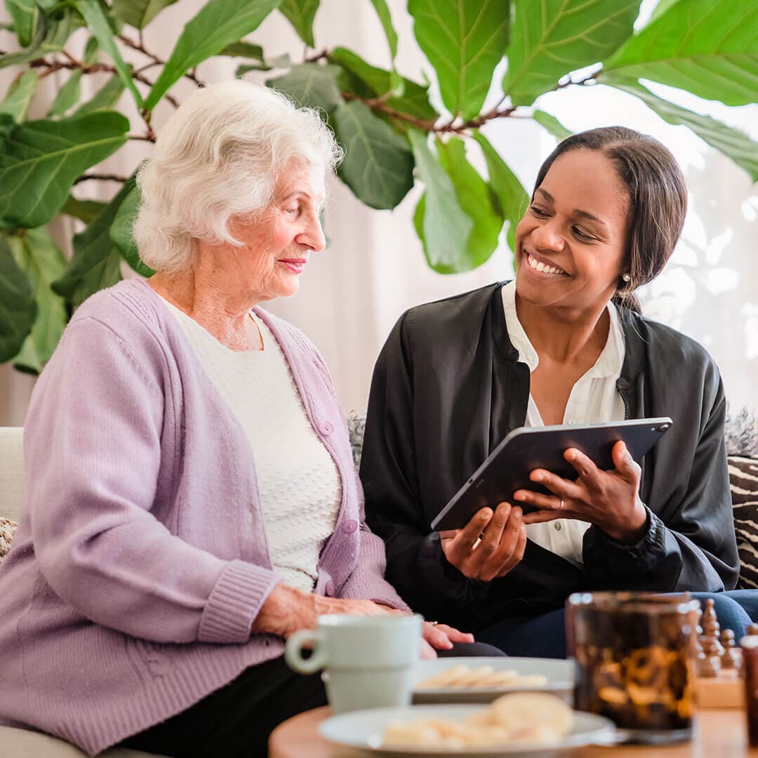 An elderly woman and a younger woman smiling and looking at a tablet together, seated in a cozy room with large leaves behind them. - Home Instead