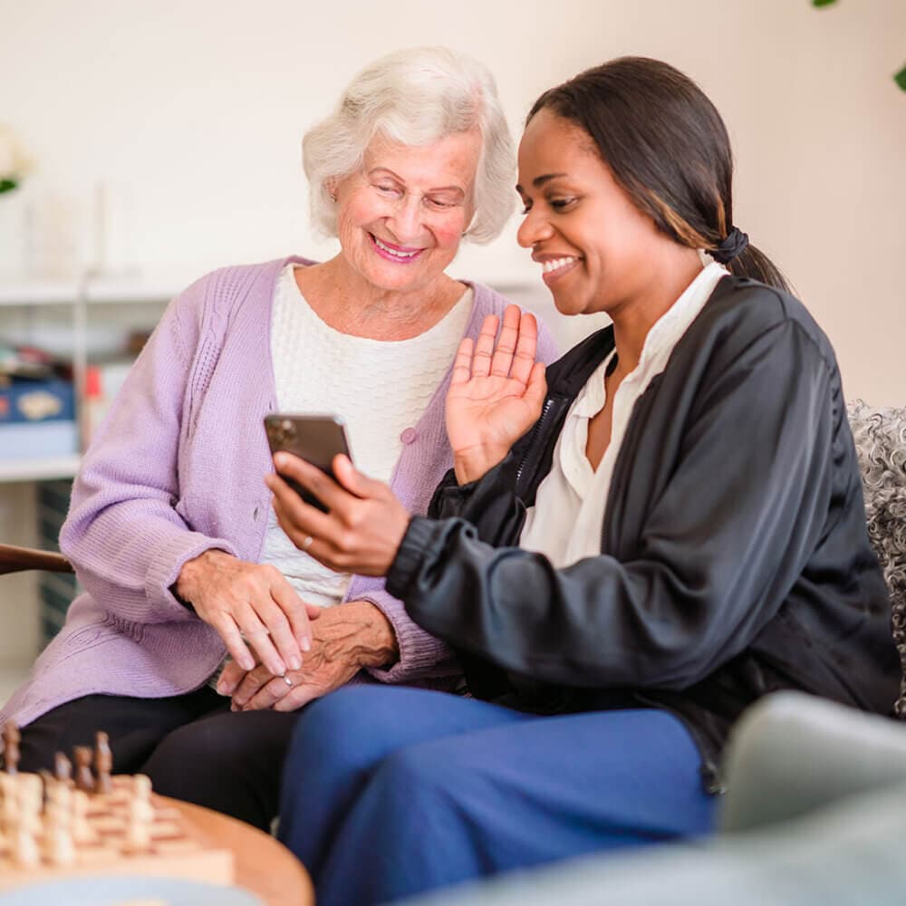Elderly woman and young woman smiling and waving at a smartphone, sitting together at a chessboard. - Home Instead