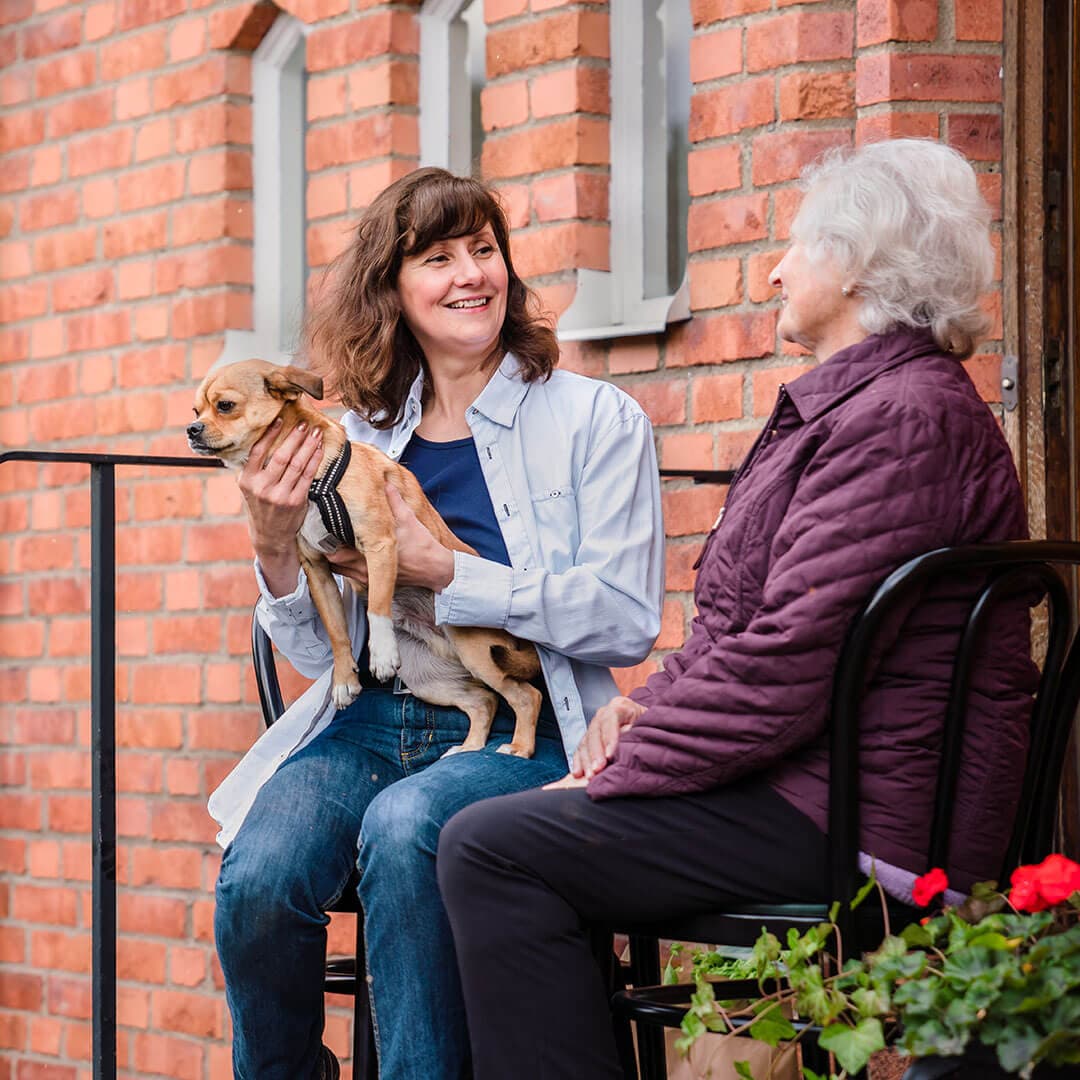 Two women, one holding a small dog, sit outside a brick building, chatting and smiling. - Home Instead