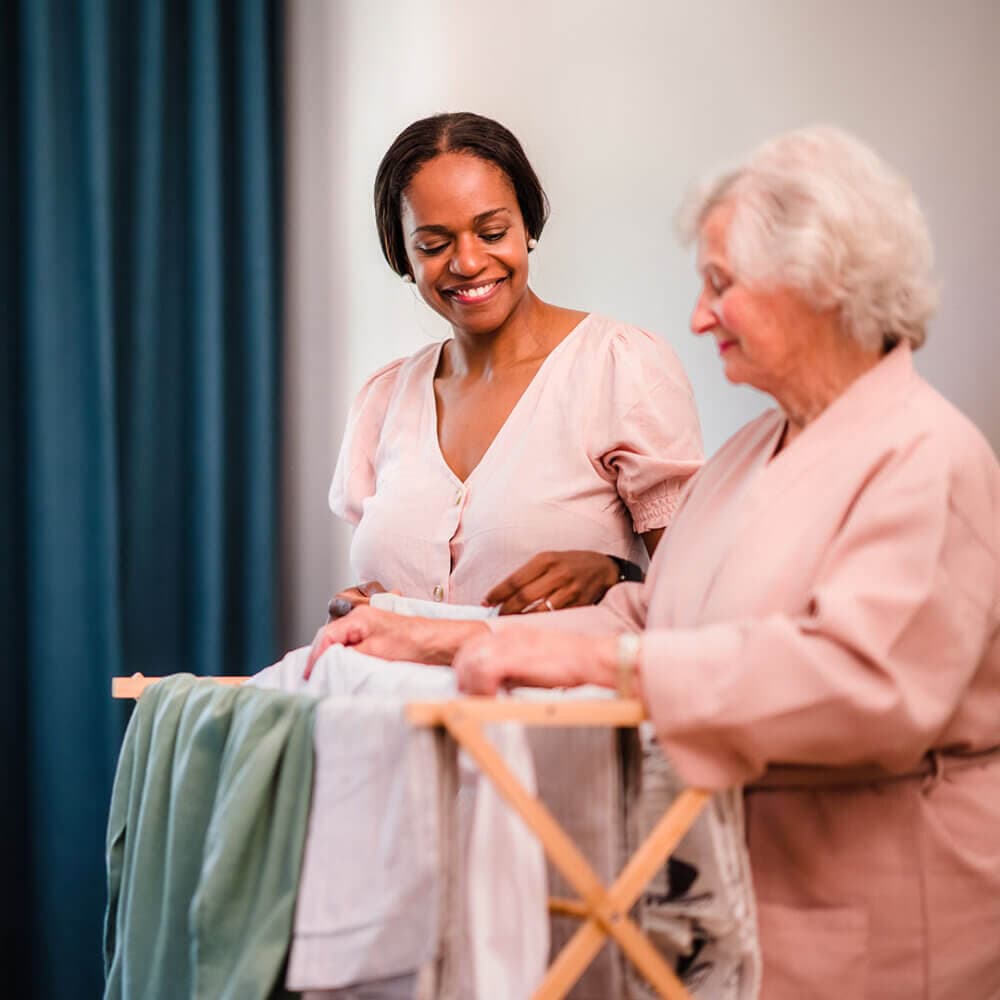 Younger woman helping older woman fold laundry indoors. Both are smiling and engaged in the activity. - Home Instead