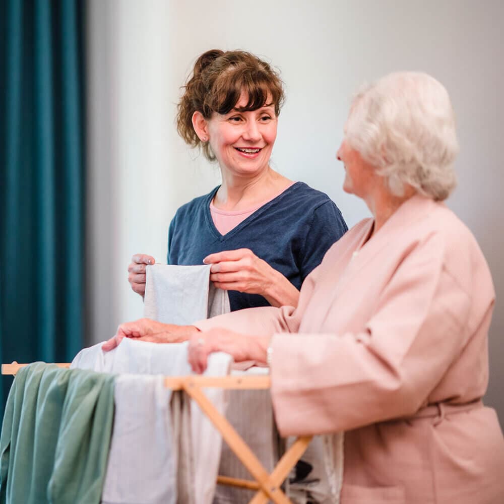 A young woman smiles at an elderly woman while they hang laundry on a drying rack together. - Home Instead