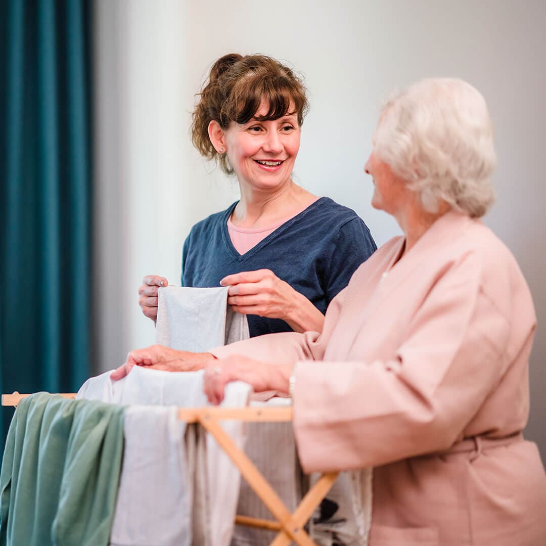 Two women, one elderly and one younger, smiling and folding laundry together near a drying rack. - Home Instead Poole
