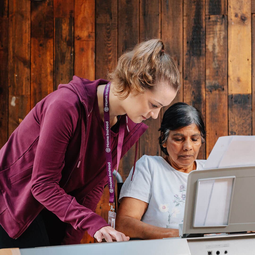 A caregiver assists an elderly woman with paperwork at a wooden table. - Home Instead