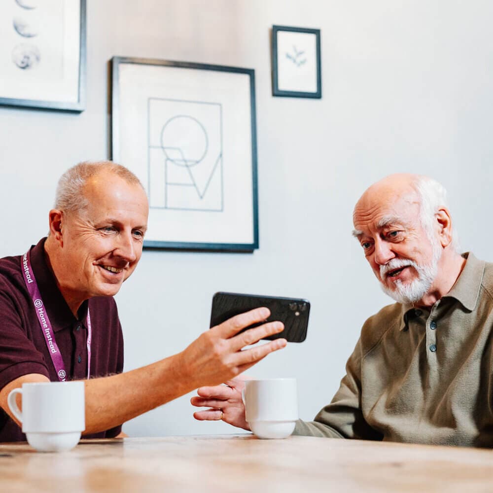 Two elderly men smiling and looking at a smartphone, seated at a wooden table with white mugs. - Home Instead