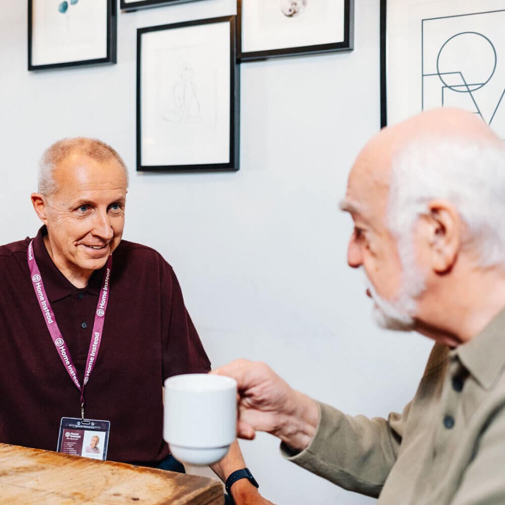 Two elderly men having a conversation at a table. One is holding a coffee cup, and the other is wearing a name tag. - Home Instead