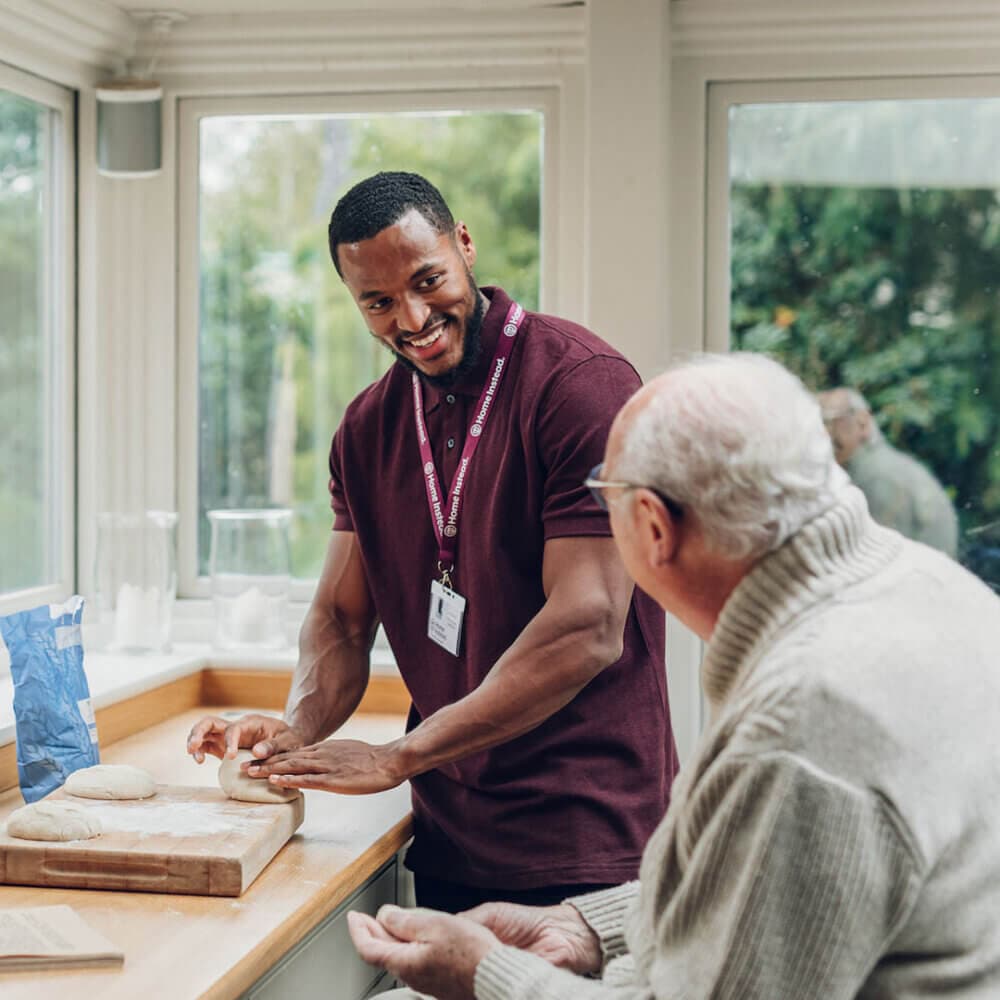A caregiver helps an elderly man in the kitchen as they prepare dough together, both smiling and engaging warmly. - Home Instead