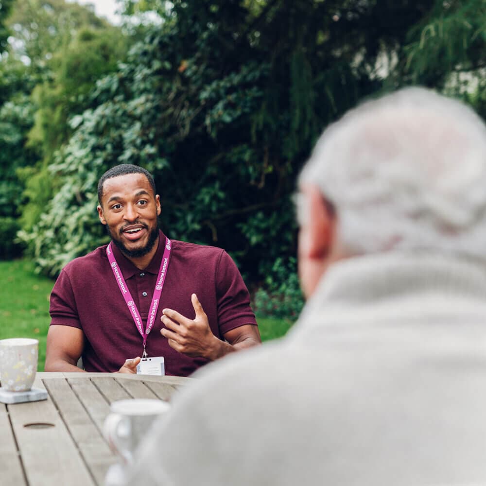 A man in a maroon shirt talks with an older person outdoors at a table with mugs, amidst a green background. - Home Instead
