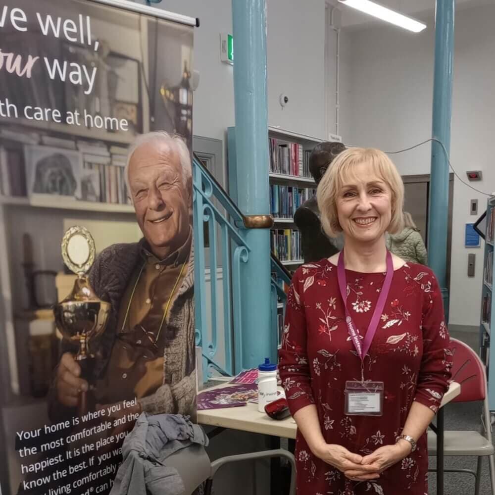 A woman stands smiling in a room beside a banner displaying an older man and text about home care services. - Home Instead