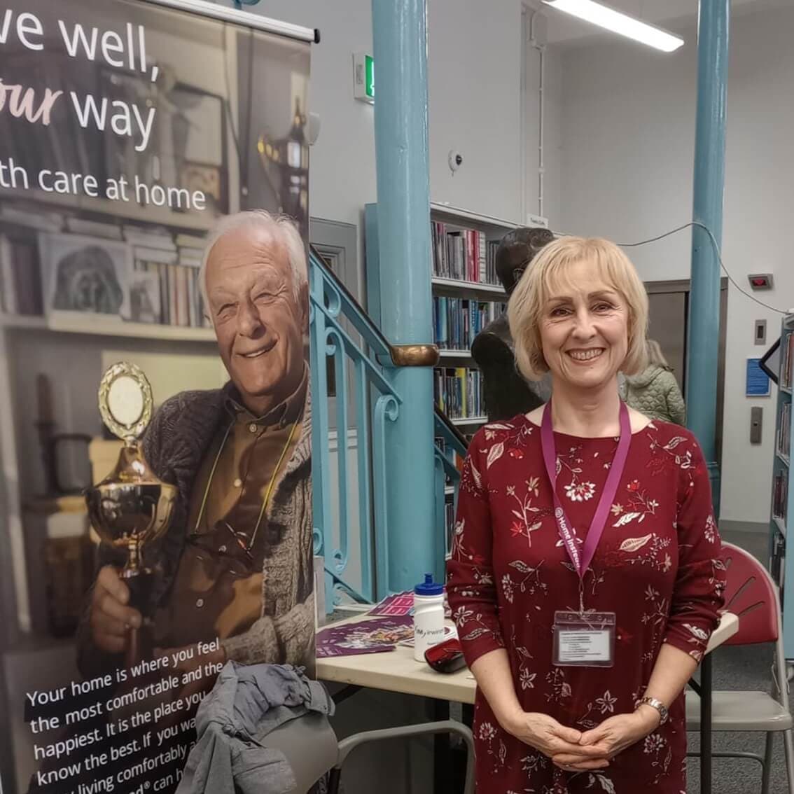 A woman stands smiling in a room beside a banner displaying an older man and text about home care services. - Home Instead