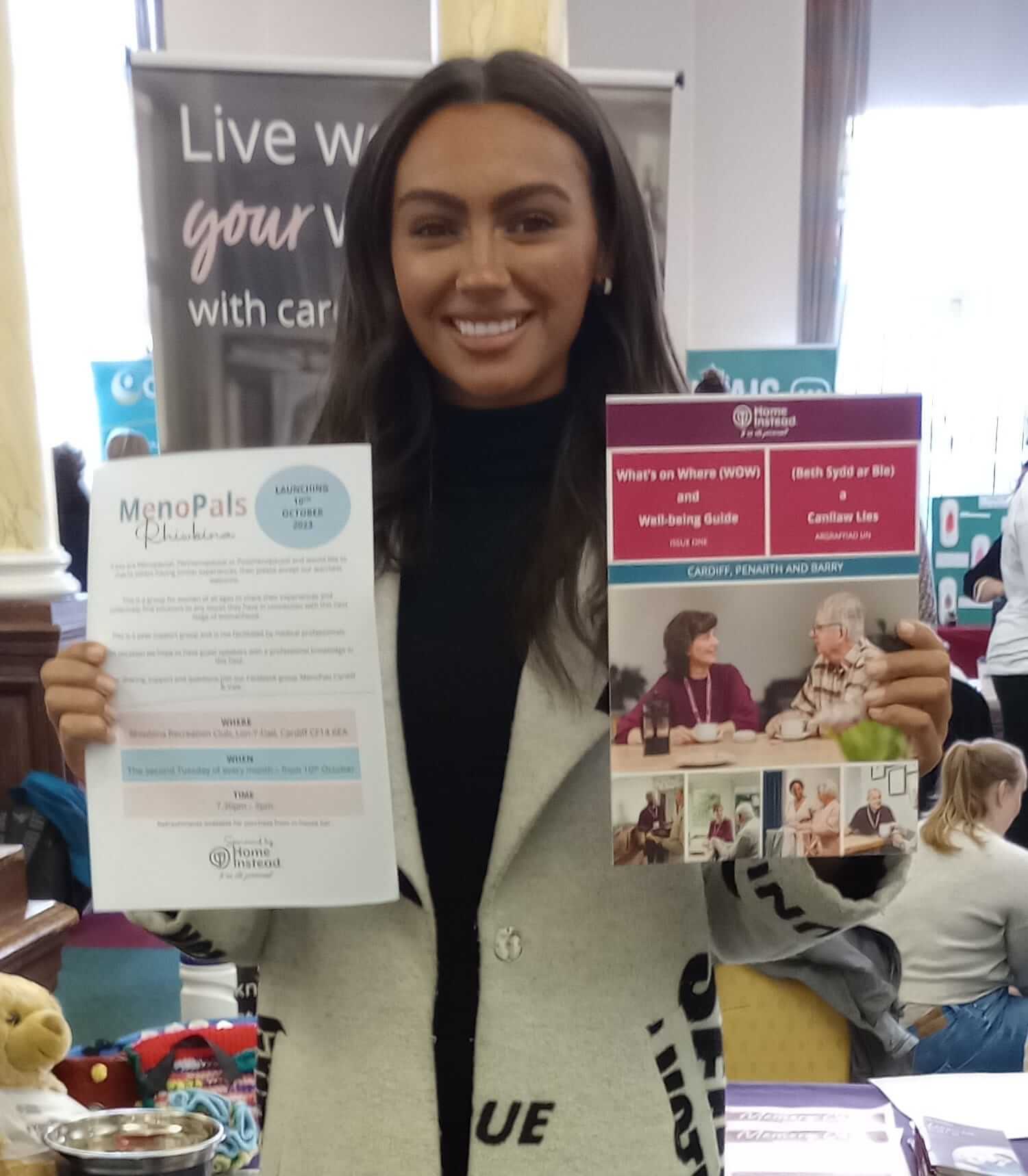 A woman smiles while holding two pamphlets at a booth with a "Live your values with carers" banner in the background. - Home Instead
