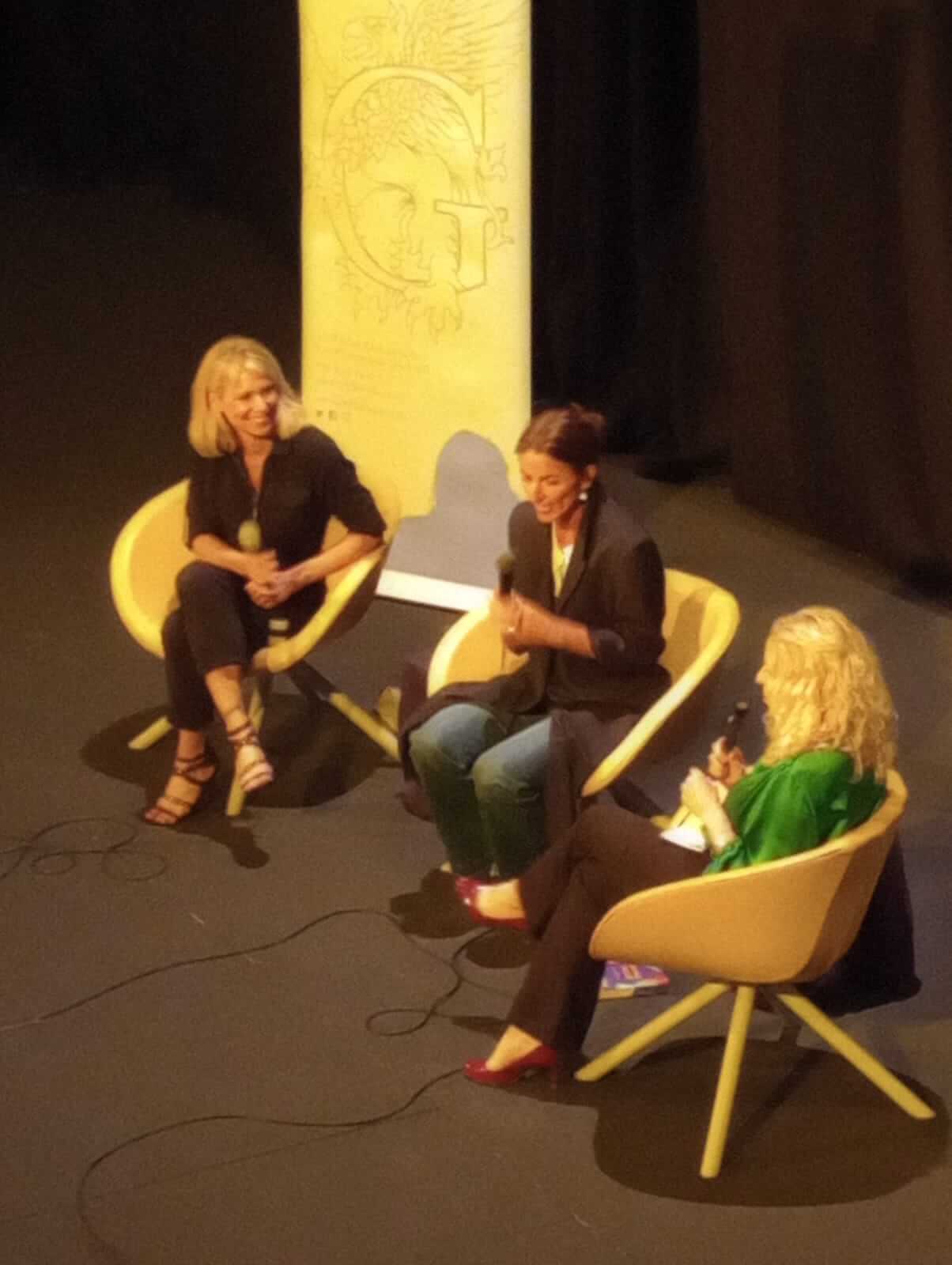 Three women sit on stage holding microphones and engaging in a discussion at an event. - Home Instead