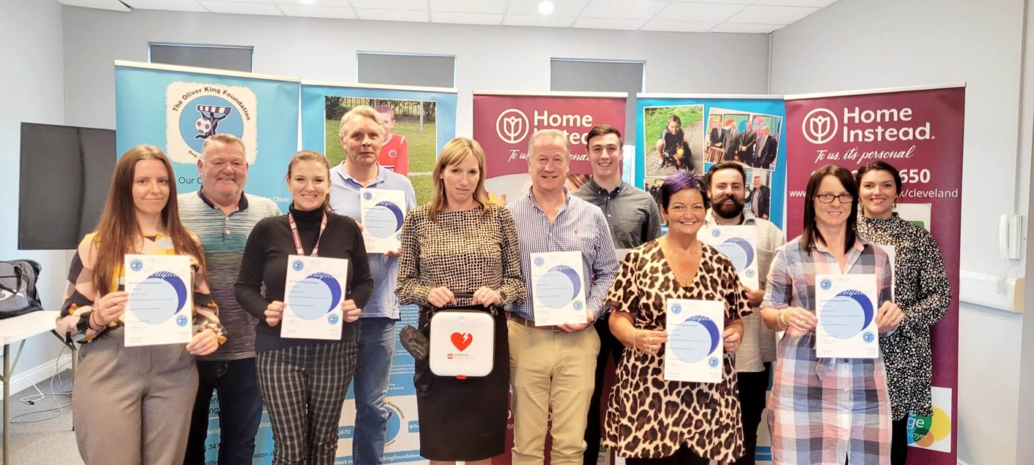 A group of people holding certificates while standing indoors in front of banners. One person holds a defibrillator. - Home Instead