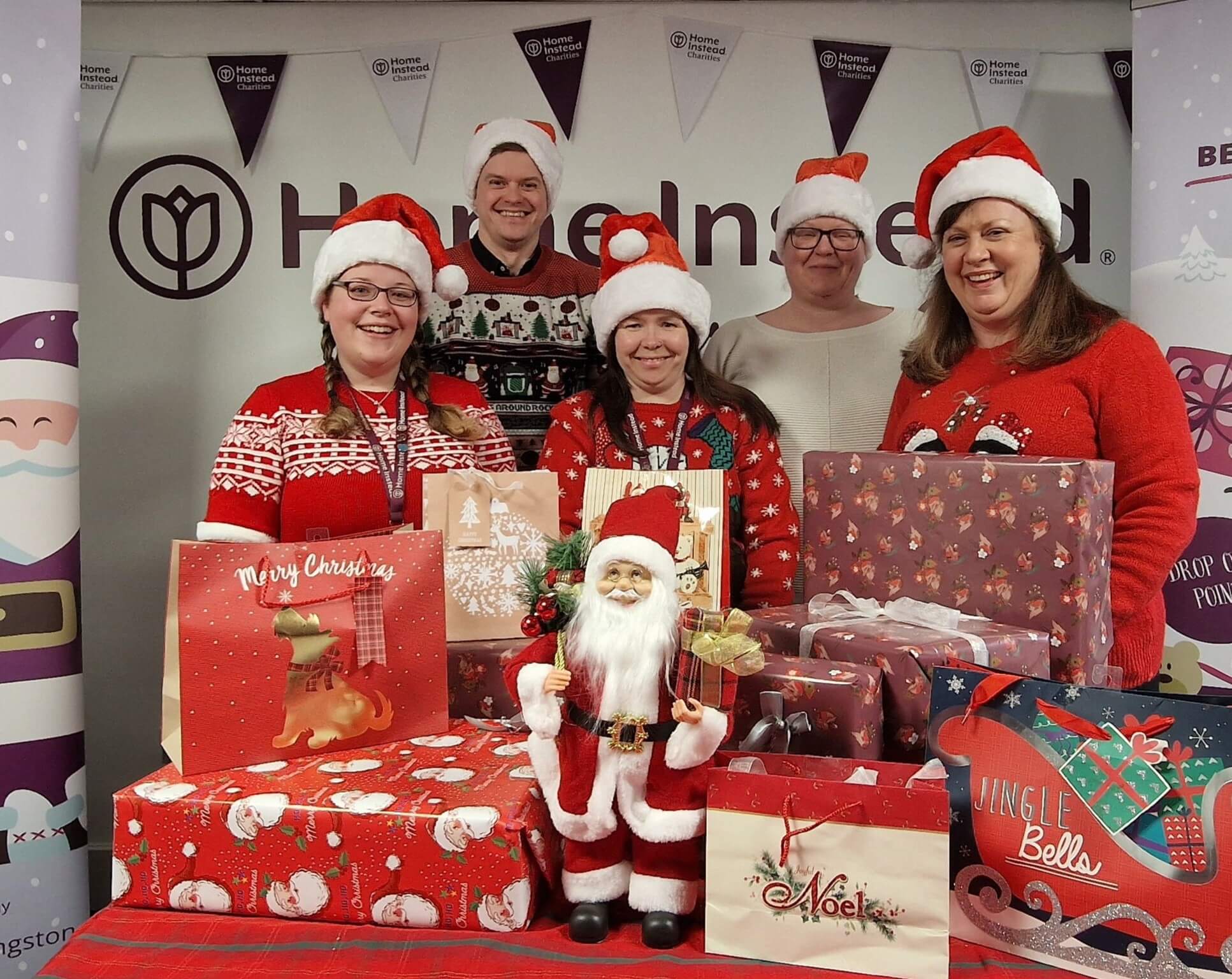 Six people in festive sweaters and Santa hats posing with wrapped Christmas gifts and a Santa Claus doll. - Home Instead