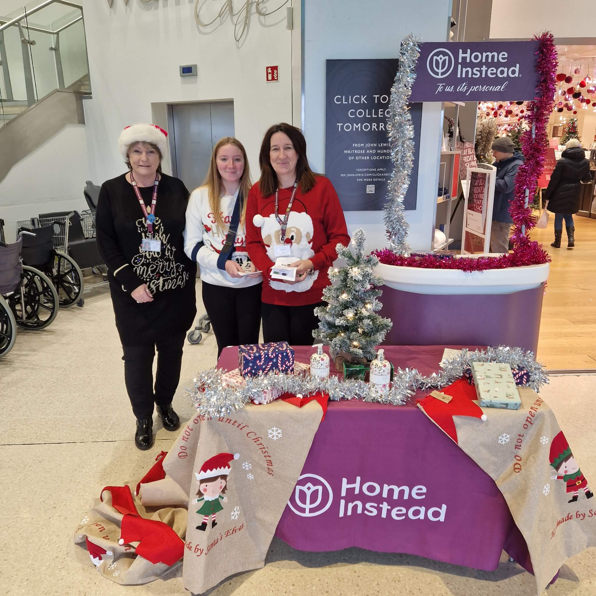 Three women at a Home Instead festive stall with a Christmas tree, brochures, and decorations in a shopping center. - Home Instead