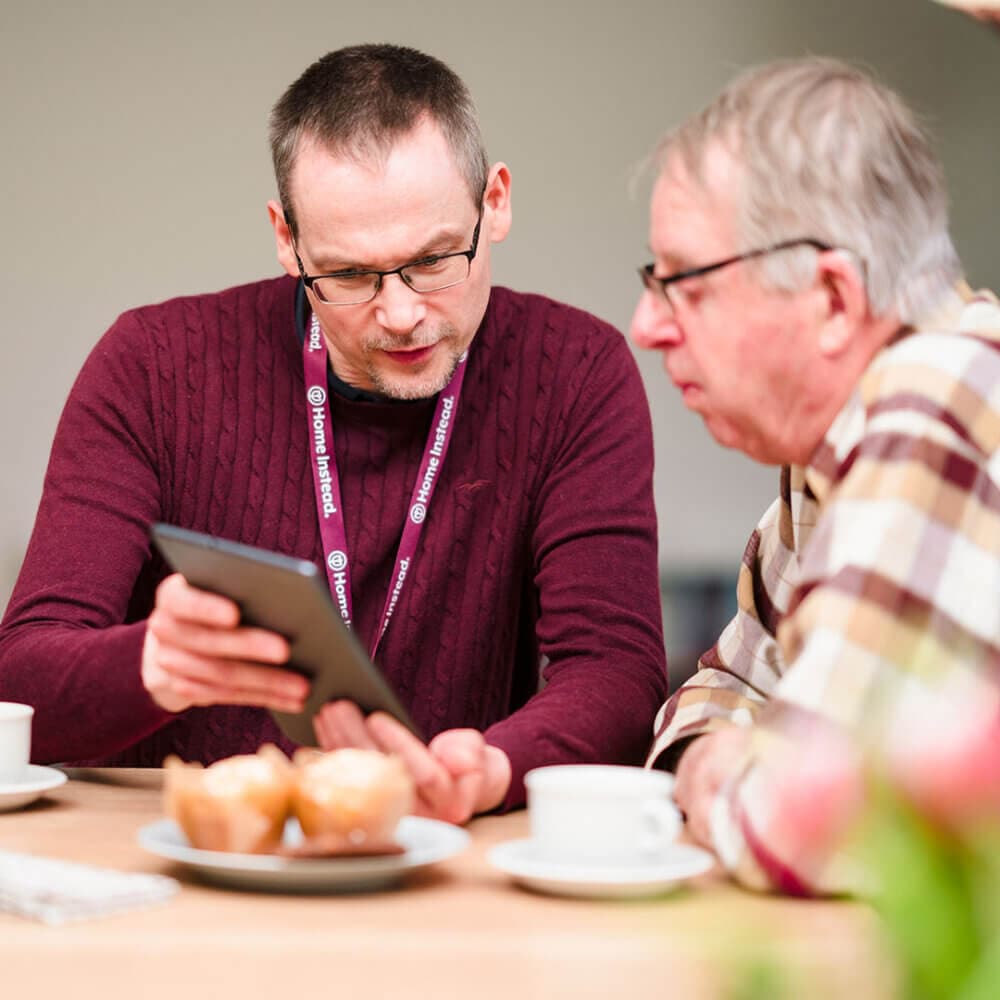 Two men sitting at a table, one showing the other something on a tablet. Cups of coffee and muffins are on the table. - Home Instead