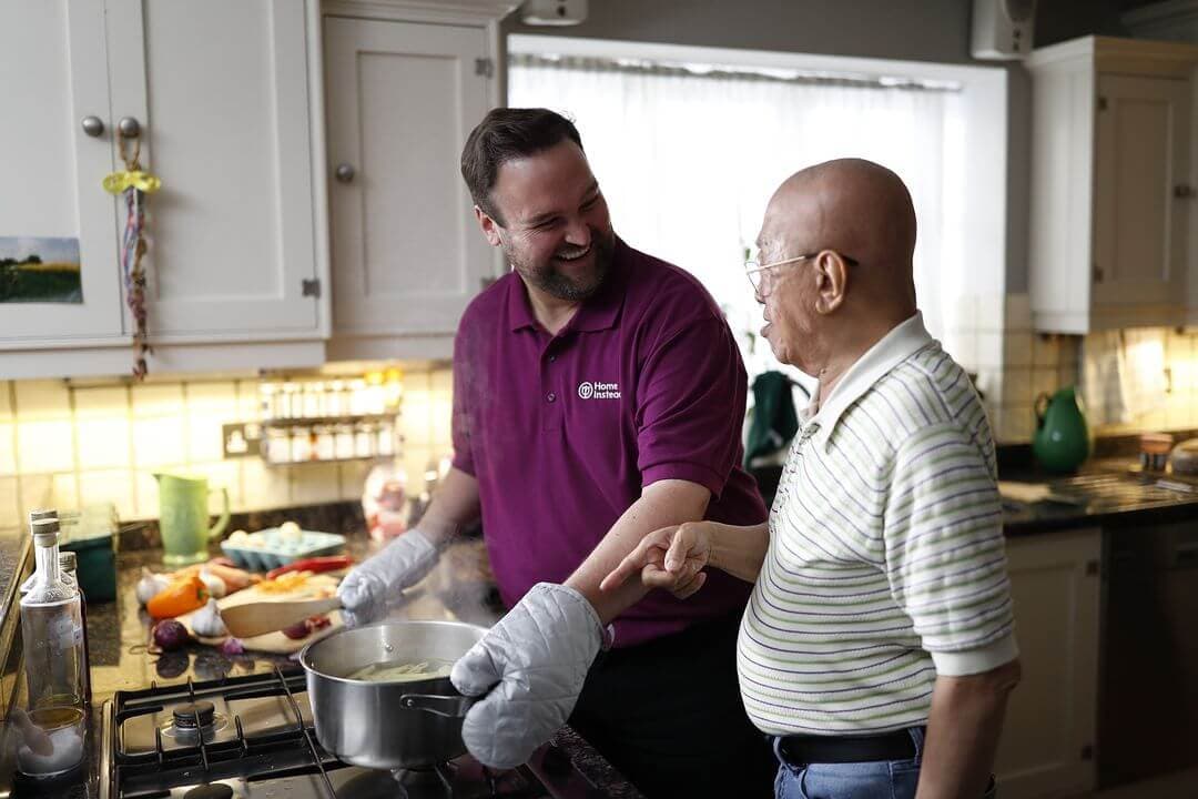 Two men in a kitchen: a caregiver wearing oven mitts helping an elderly man cook at the stovetop. Both are smiling. - Home Instead