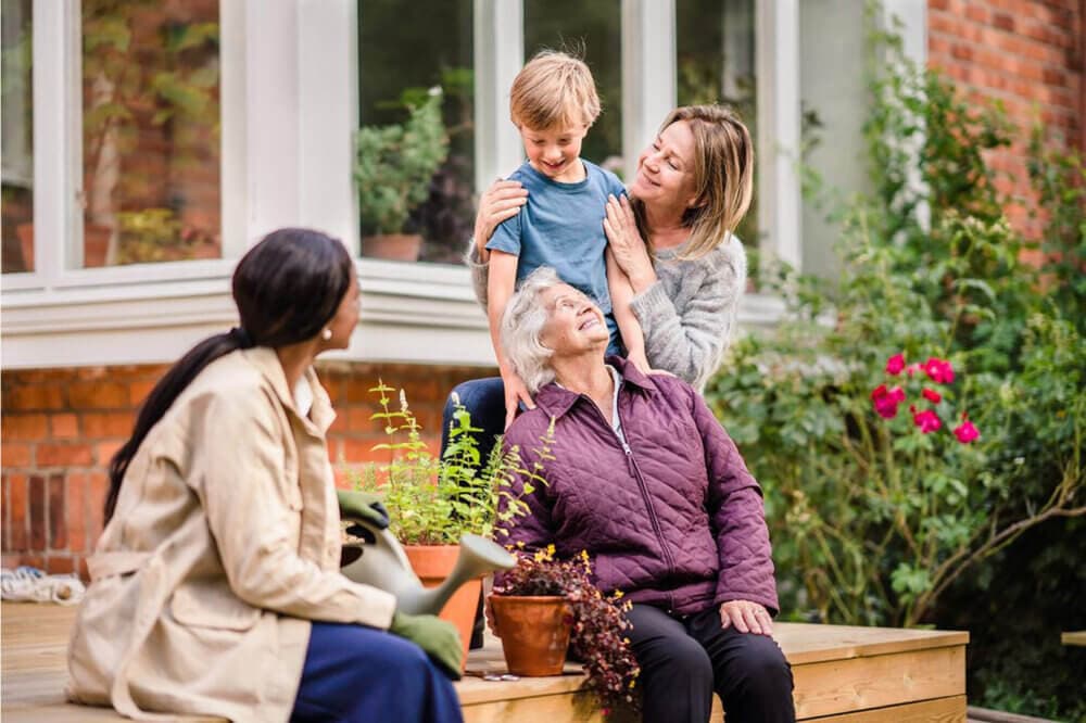 A family with three women and a young boy smiles and interacts on a wooden deck surrounded by plants and flowers. - Home Instead