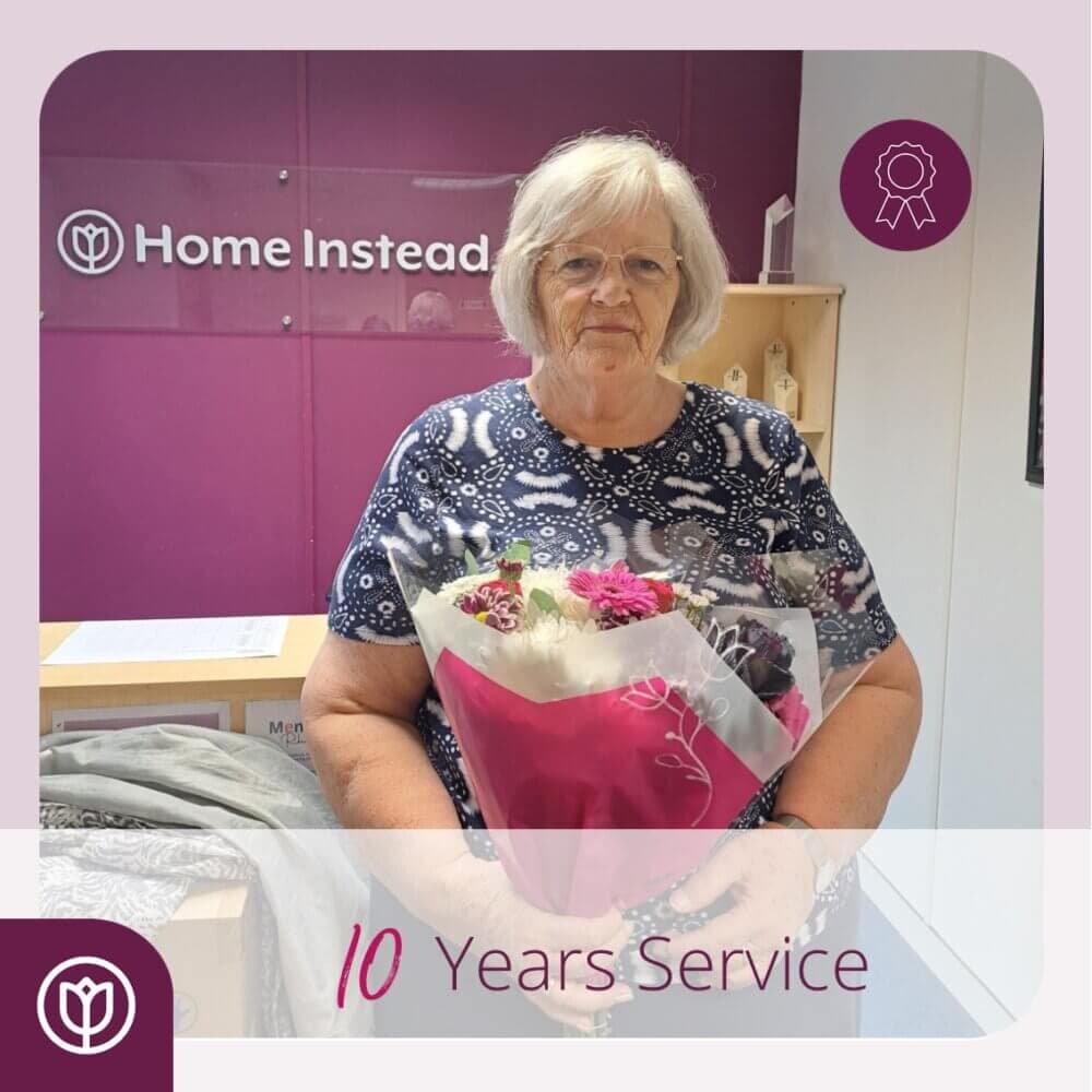 A woman holding a bouquet stands in front of a "Home Instead" sign, celebrating 10 years of service. - Home Instead