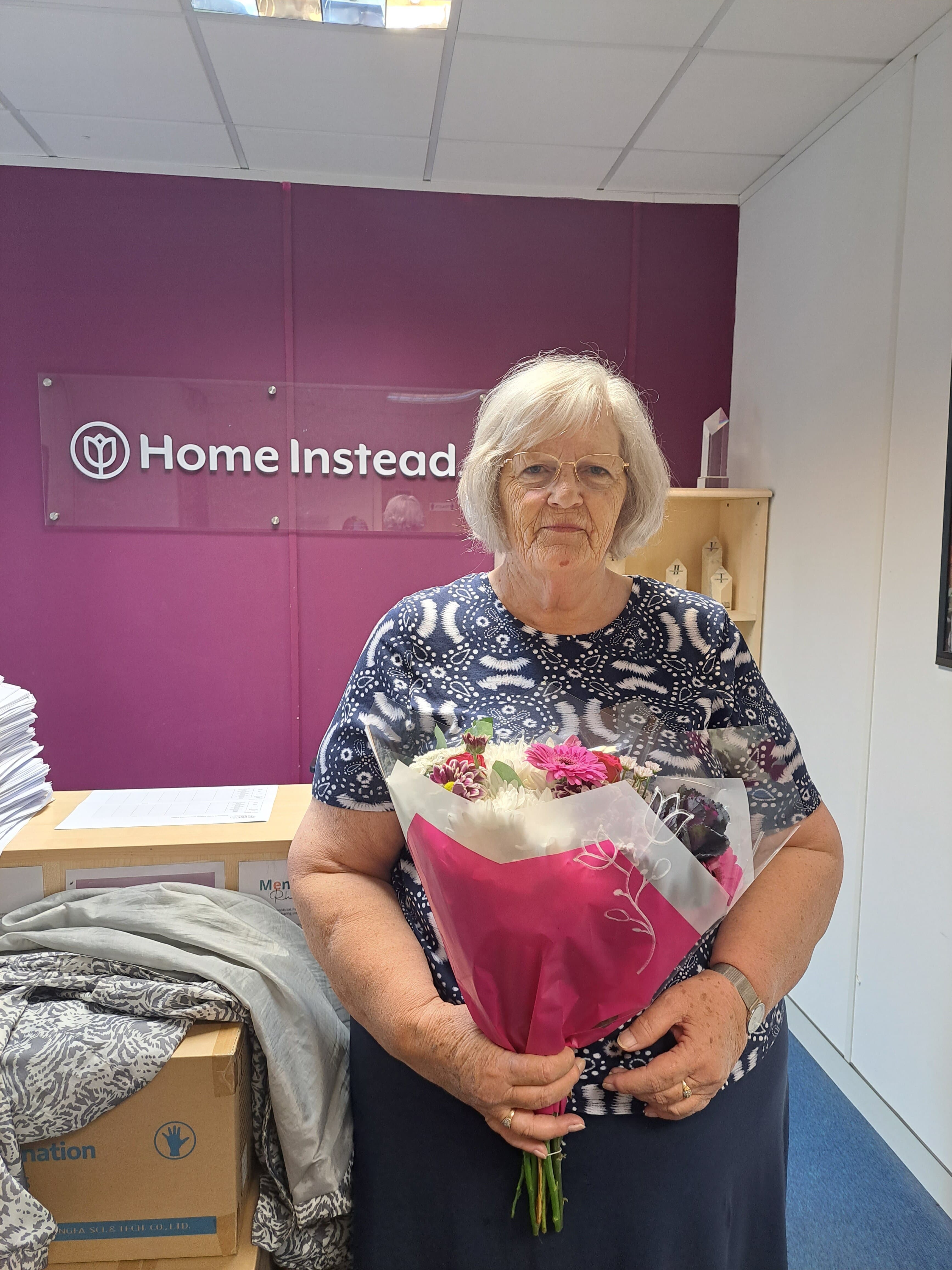 An elderly woman stands holding a bouquet of flowers in an office with a "Home Instead" sign in the background. - Home Instead