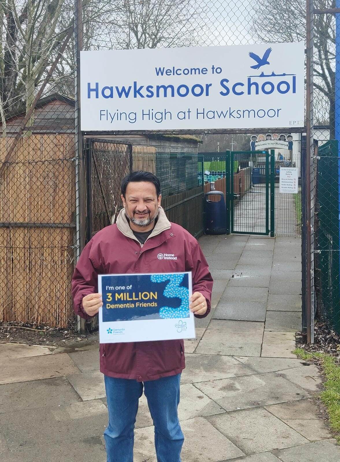 A man holds a sign outside Hawksmoor School that reads "I'm one of 3 MILLION Dementia Friends," smiling at the camera. - Home Instead