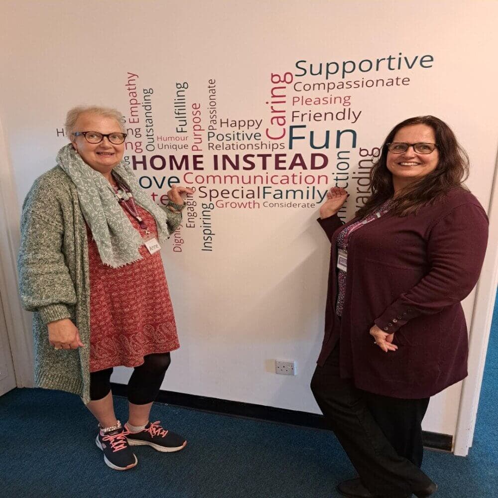 Two smiling women stand beside a word cloud wall art, with one woman pointing at it. Both are wearing lanyards. - Home Instead
