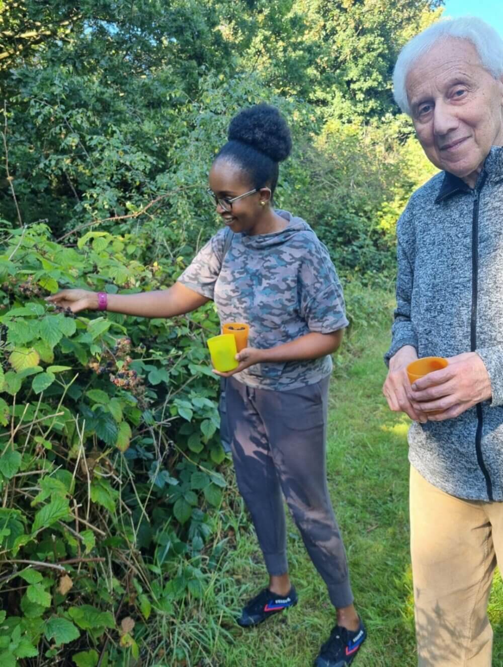 A woman and an elderly man gather berries outdoors, holding cups and standing on a grassy path surrounded by greenery. - Home Instead
