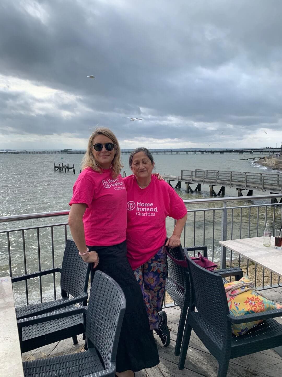 Two women in pink "Home Instead Charities" t-shirts smiling by the waterfront on a cloudy day. - Home Instead